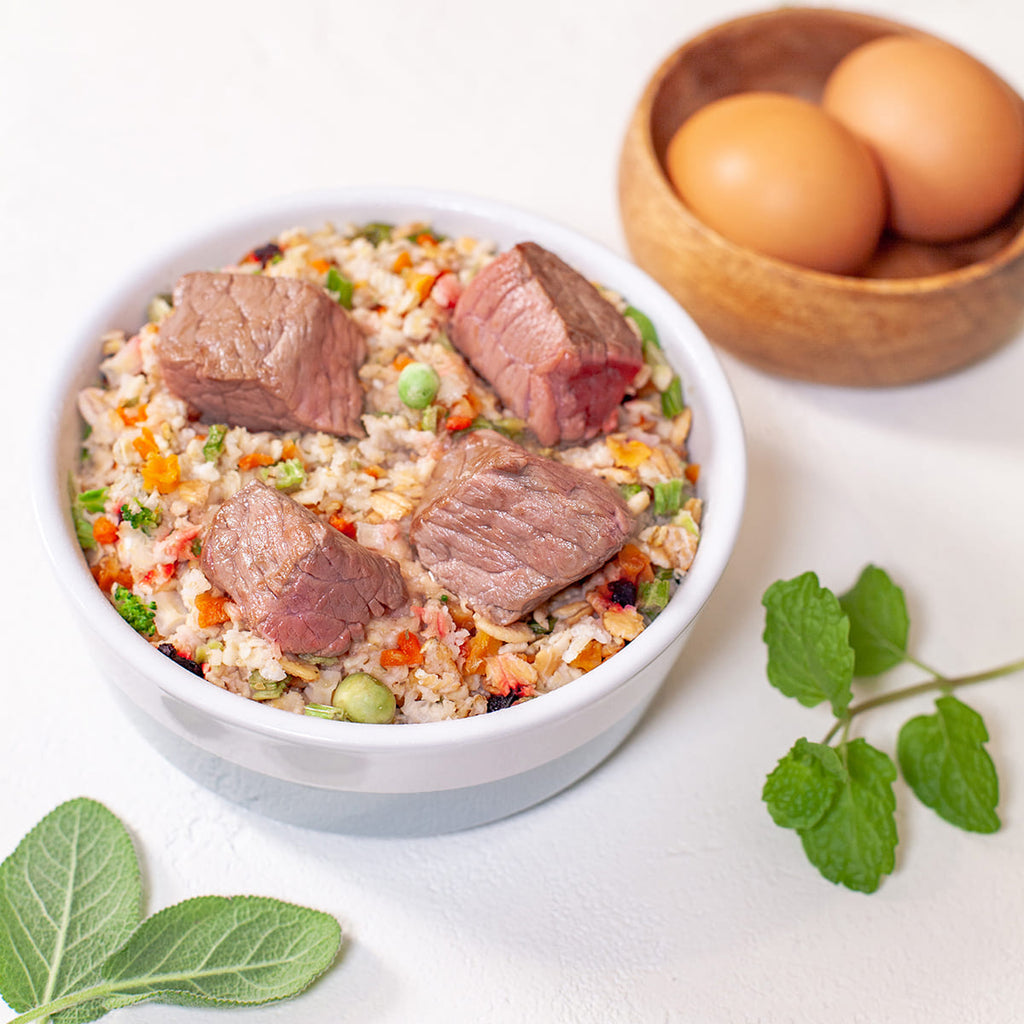 White bowl of beef and vegetable rice topped with meat chunks, beside eggs in a wooden bowl and fresh herbs.