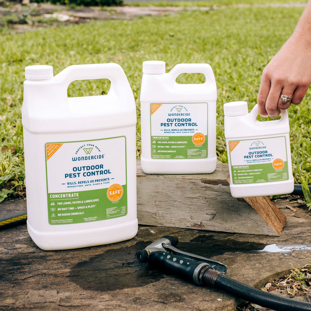 Three Wondercide Outdoor Pest Control Concentrate for Yard + Garden containers are on a wooden plank outside with grass in the background. A hand holds the smallest, and a garden hose nozzle—perfect for applying this mosquito repellent—is nearby.