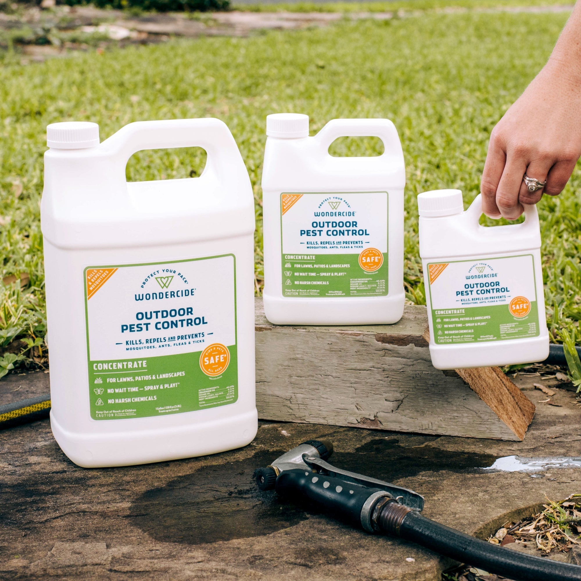 Three Wondercide Outdoor Pest Control Concentrate for Yard + Garden containers are on a wooden plank outside with grass in the background. A hand holds the smallest, and a garden hose nozzle—perfect for applying this mosquito repellent—is nearby.