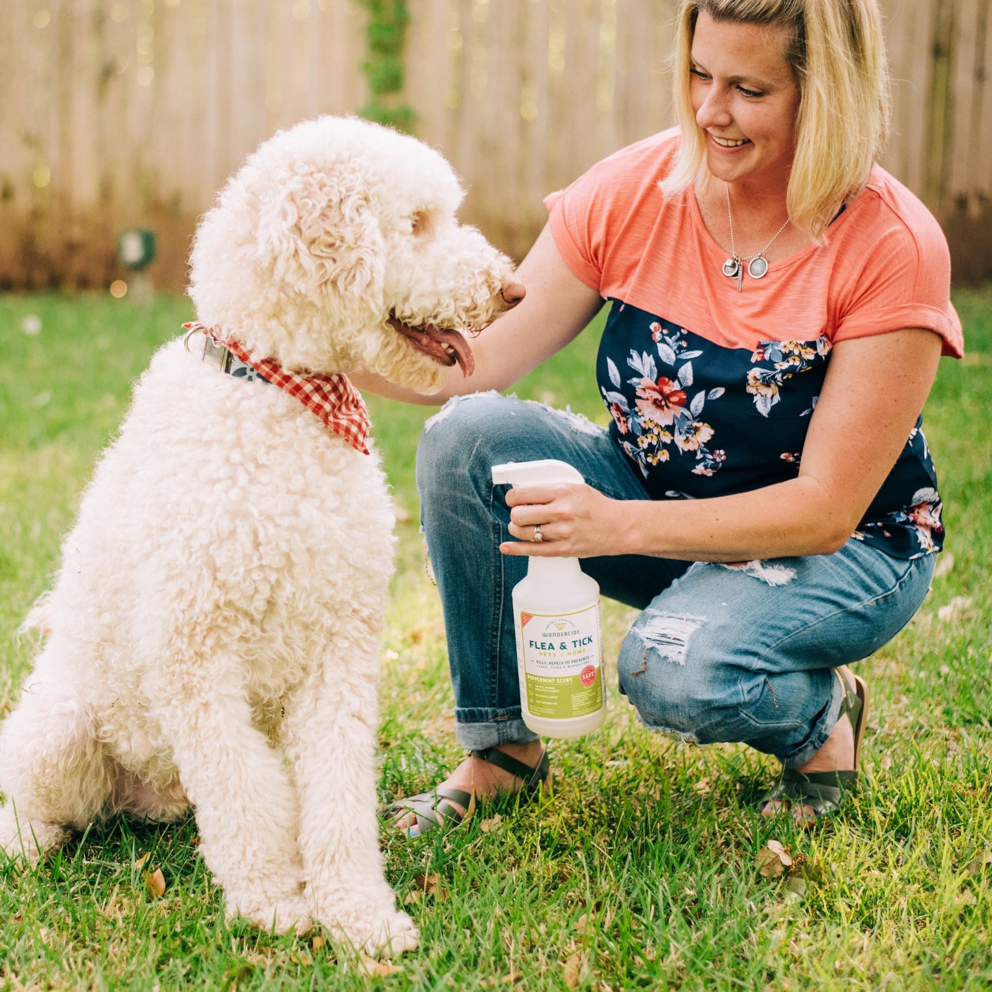 A woman kneels on grass in her backyard, smiling at a large white dog in a red bandana while holding Wondercide Flea & Tick Spray for Pets + Home. A wooden fence is visible in the background.