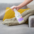 A hand sprays Wondercide Flea & Tick Spray for Pets + Home onto a gray couch with a green blanket and yellow textured pillow in the background, safe for both dogs and cats.