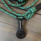 Image shows a teal and black climbing rope-style dog leash from Wilderdog, coiled with a plastic loop holder attached. The leash, laid out on a wooden surface, showcases its durable weave and the brand name prominently displayed on the holder. A hands-free Crap Carrier attachment ensures convenience during walks.