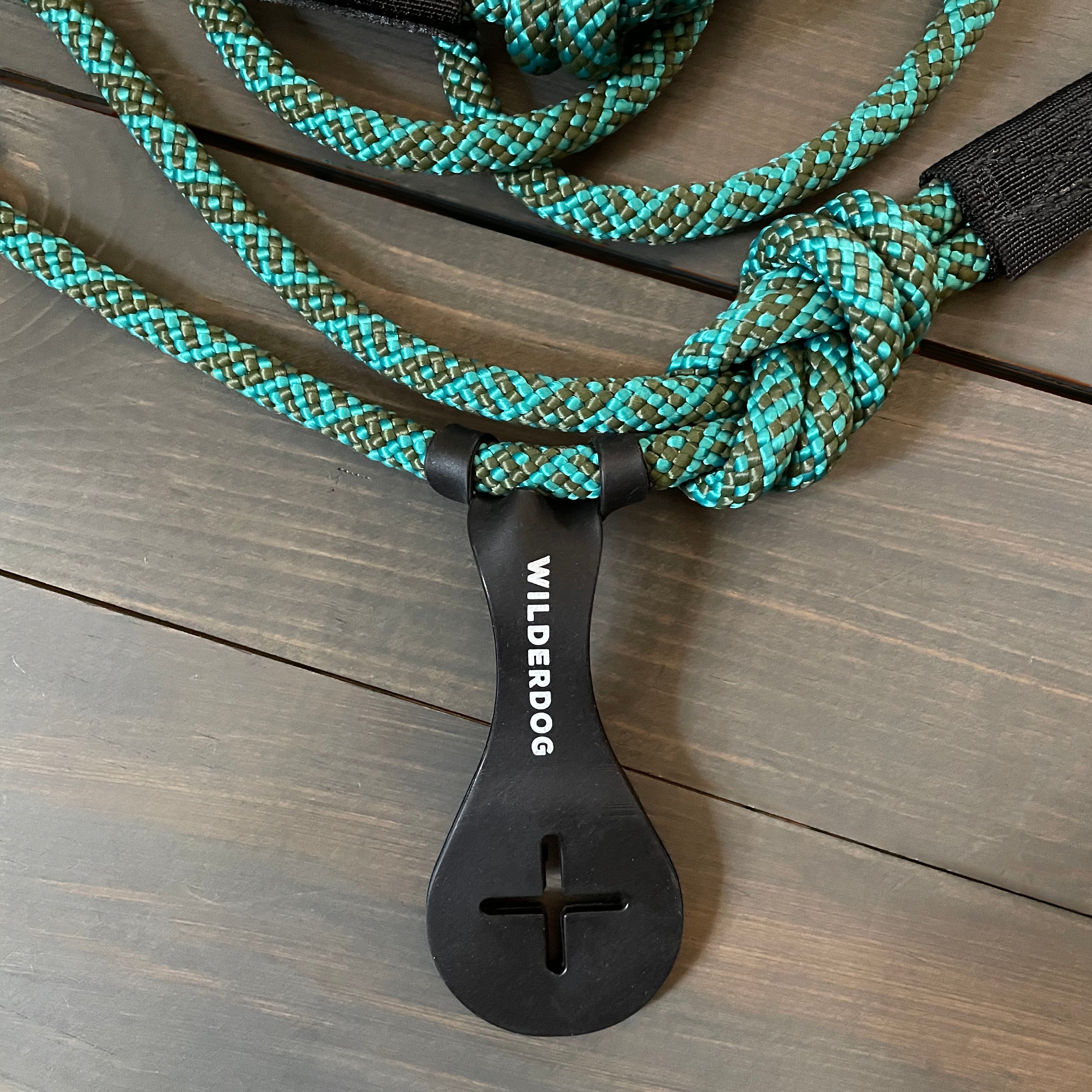 Image shows a teal and black climbing rope-style dog leash from Wilderdog, coiled with a plastic loop holder attached. The leash, laid out on a wooden surface, showcases its durable weave and the brand name prominently displayed on the holder. A hands-free Crap Carrier attachment ensures convenience during walks.