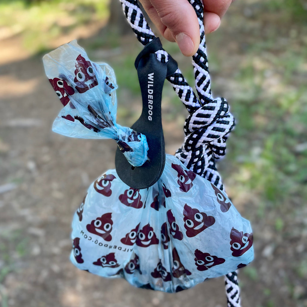 A hand holding a black and white rope with a blue Crap Carrier dog waste bag attached. The waste bag, decorated with poop emojis and tied in a knot at the top, serves as an efficient dog poop carrier. Connected to the rope with a Wilderdog brand carabiner, it offers hands-free convenience in an outdoor setting.
