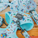 A close-up of the Dog Adjustable Harness - Disney's Stitch by Sassy Woof in light blue, featuring colorful Stitch, palm trees, and surfboards. The adjustable harness has a Sassy Woof label and the Lilo & Stitch logo in the corner.
