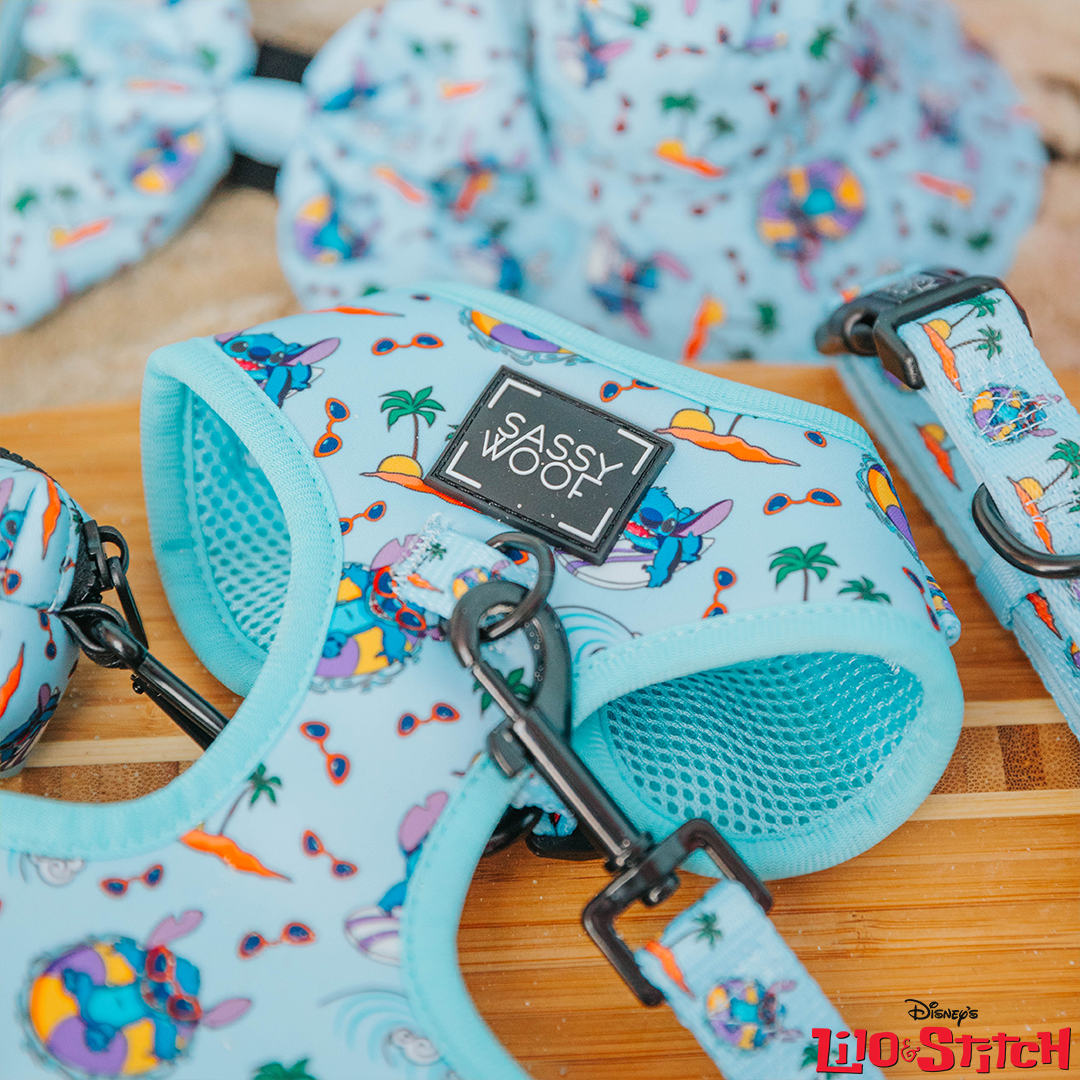 A close-up of the Dog Adjustable Harness - Disney's Stitch by Sassy Woof in light blue, featuring colorful Stitch, palm trees, and surfboards. The adjustable harness has a Sassy Woof label and the Lilo & Stitch logo in the corner.