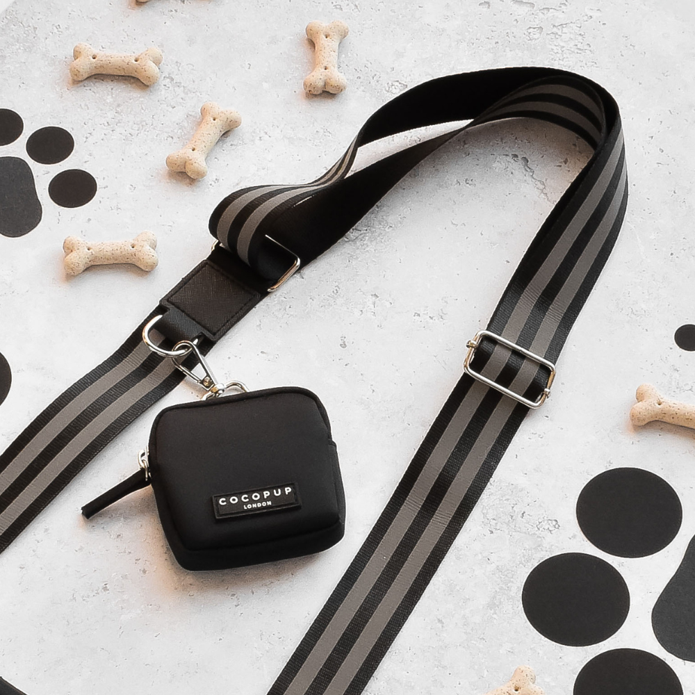 Black dog walking bag by Cocopup London with striped leash, treat pouch, dog bones, and paw prints placed nearby.