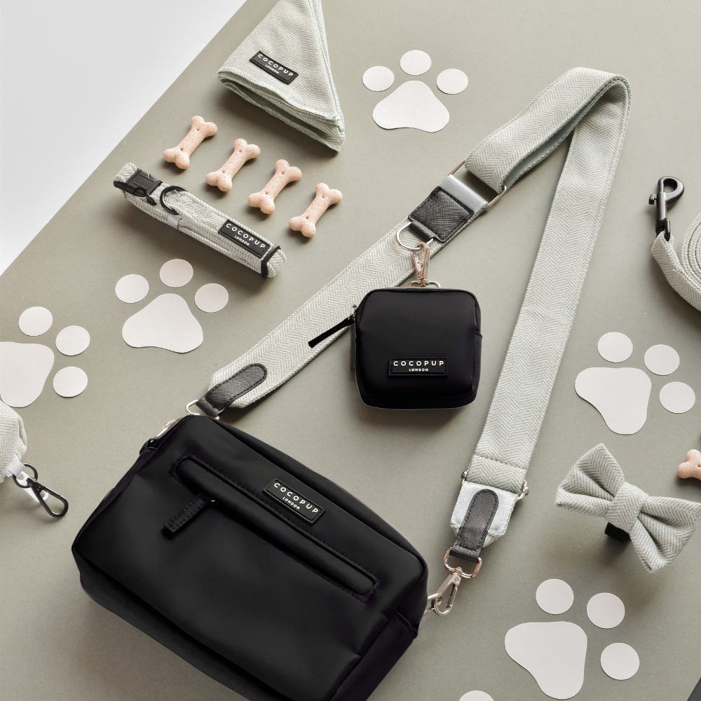 Black Cocopup London dog walking bag, matching leash, collar, treat pouch, bow tie and toy bones on grey background.