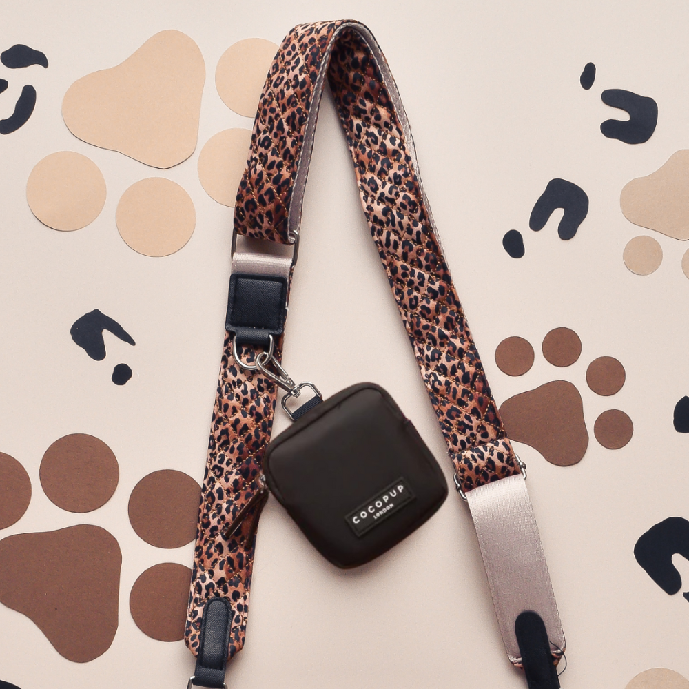 Black Cocopup London dog walking bag with a leopard-print leash, shown on a light surface with paw prints.