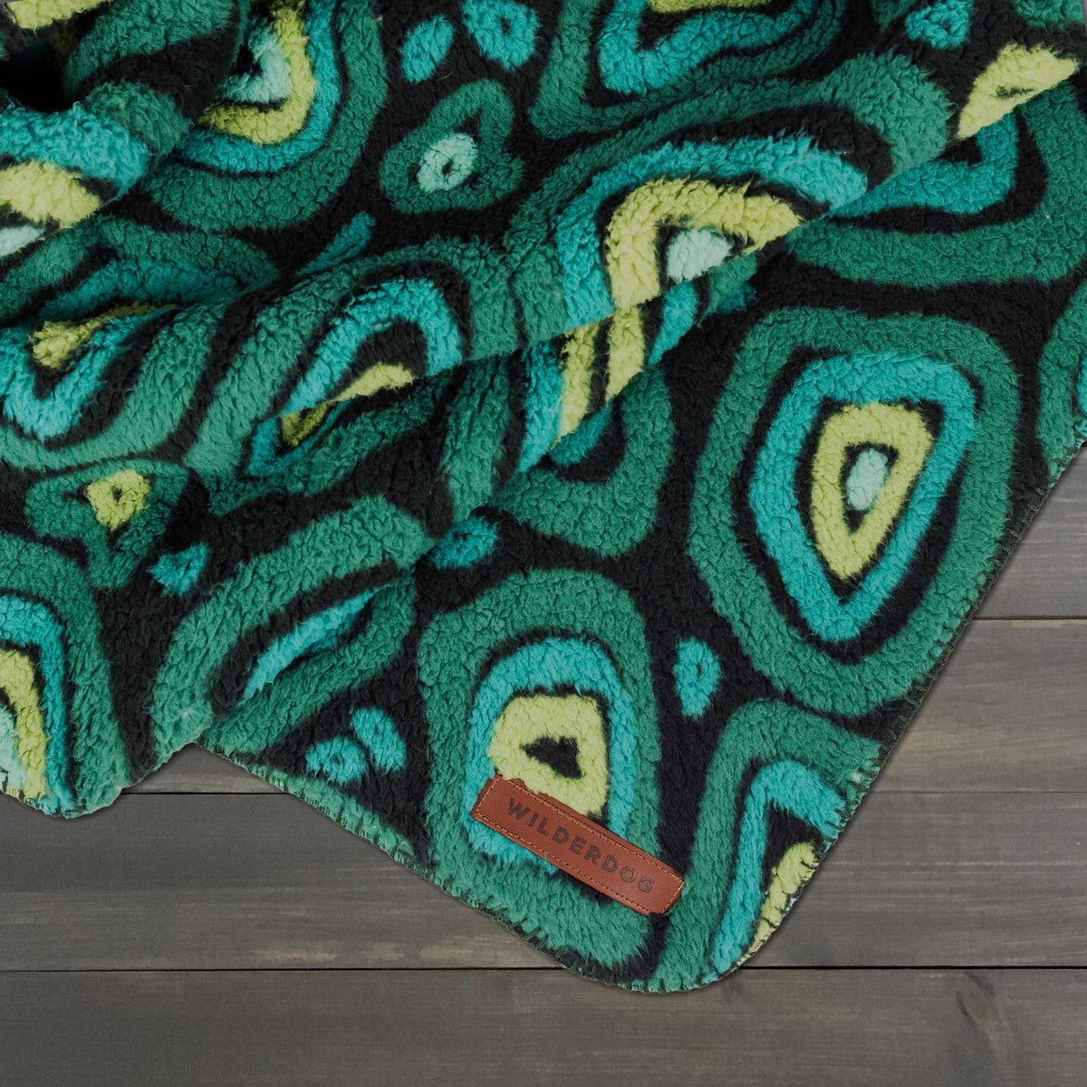 Green, blue, and yellow Sherpa Fleece Waterproof Blanket with label, shown on a dark wood surface.