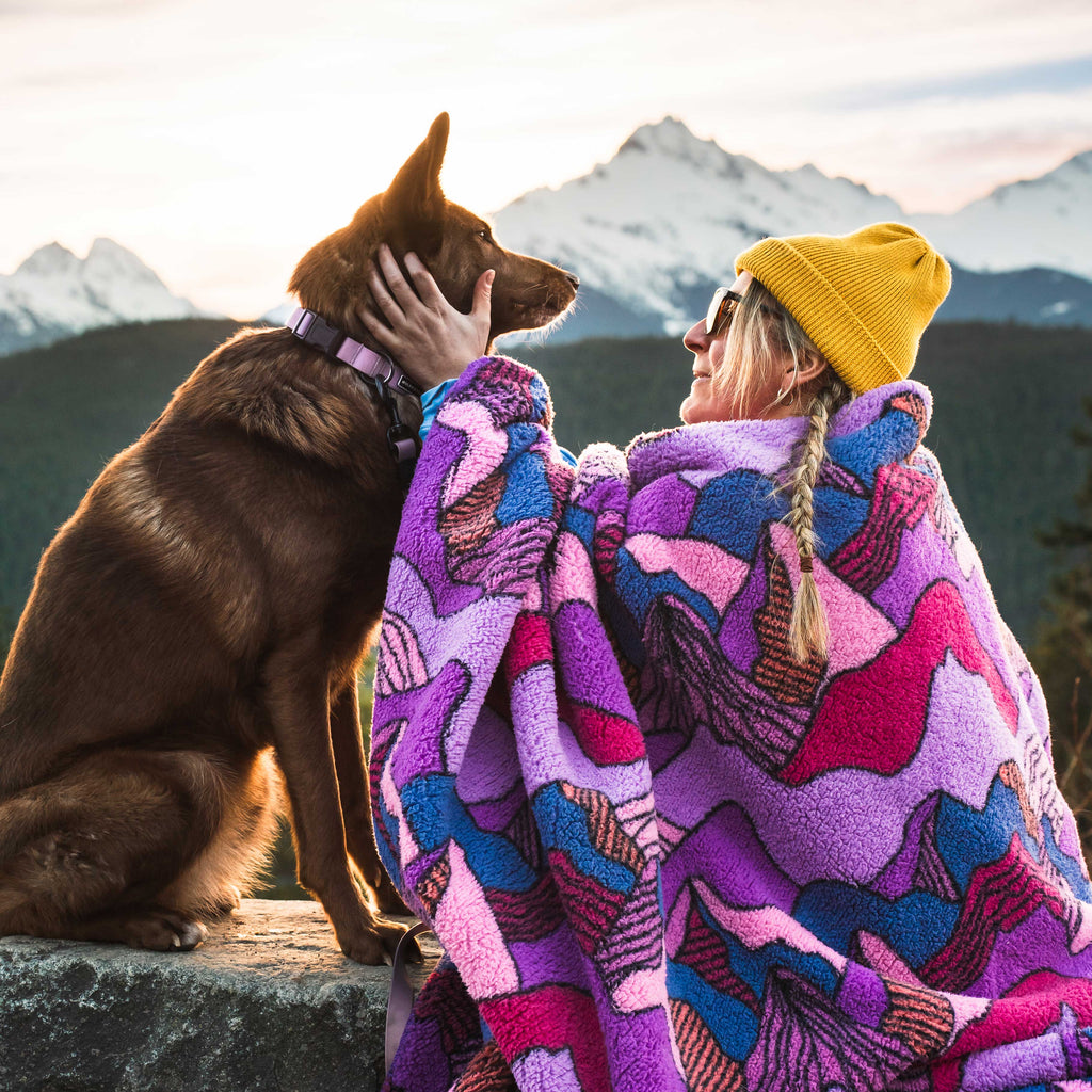 A person in a yellow beanie and sunglasses sits on a stone ledge at sunset, gently holding a brown dog’s face while wrapped in a Sherpa Fleece Waterproof Blanket. Snowy mountains and forested hills are visible in the background.