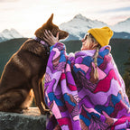 A person in a yellow beanie and sunglasses sits on a stone ledge at sunset, gently holding a brown dog’s face while wrapped in a Sherpa Fleece Waterproof Blanket. Snowy mountains and forested hills are visible in the background.