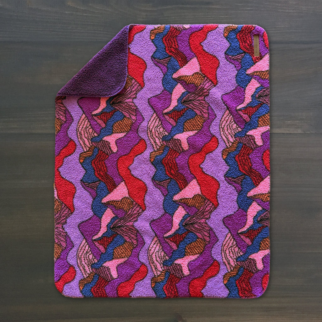 A Sherpa Fleece Waterproof Blanket with one corner folded over, displaying a vibrant abstract pattern in purple, red, pink, and orange on a dark wooden surface.