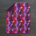 A Sherpa Fleece Waterproof Blanket with one corner folded over, displaying a vibrant abstract pattern in purple, red, pink, and orange on a dark wooden surface.