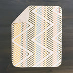 A Sherpa Fleece Waterproof Blanket, featuring a zigzag pattern in yellow, blue, pink, and brown on a white background with one corner flipped, lays on a dark wooden surface—evoking cozy bath mat style.