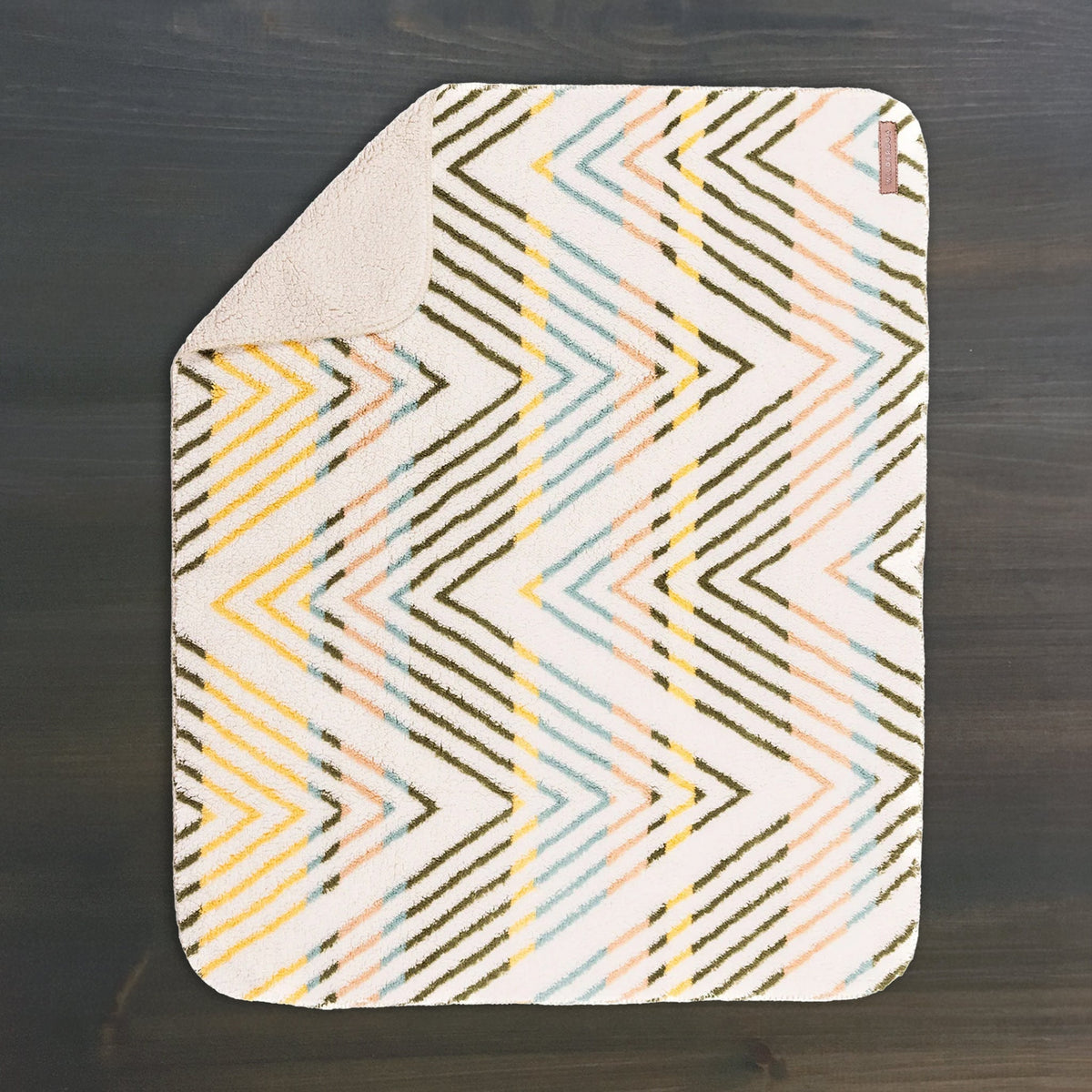 A Sherpa Fleece Waterproof Blanket, featuring a zigzag pattern in yellow, blue, pink, and brown on a white background with one corner flipped, lays on a dark wooden surface—evoking cozy bath mat style.