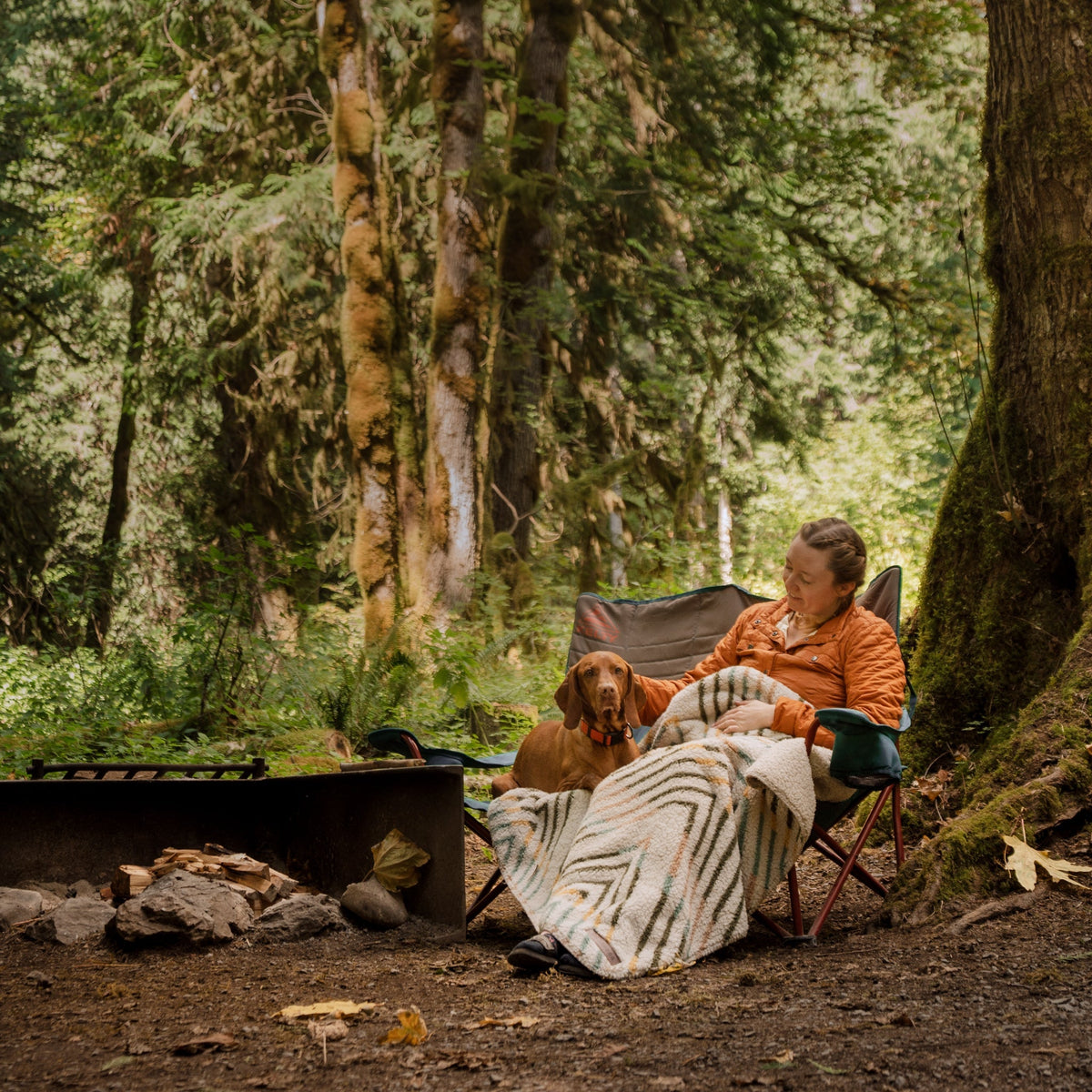 A person sits in a camping chair wrapped in a Sherpa Fleece Waterproof Blanket while petting a dog. They are in a forested area with lush green trees, and there's a portable fire pit with rocks in the foreground. The scene is peaceful and serene, evoking a cozy camping atmosphere.