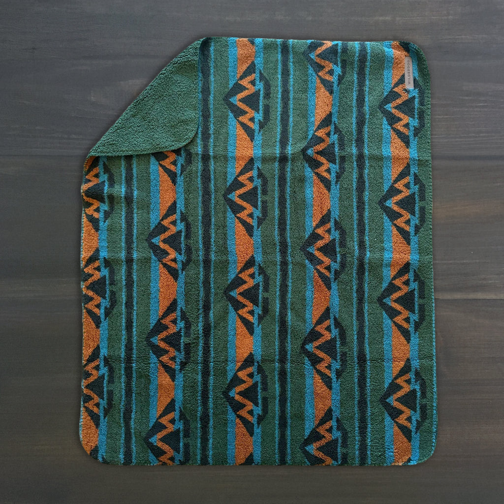 The Sherpa Fleece Waterproof Blanket, featuring a teal, green, orange, and black geometric pattern, rests on a dark wooden surface with one corner folded to show its solid green underside.