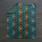 The Sherpa Fleece Waterproof Blanket, featuring a teal, green, orange, and black geometric pattern, rests on a dark wooden surface with one corner folded to show its solid green underside.
