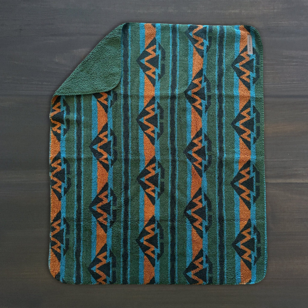 The Sherpa Fleece Waterproof Blanket, featuring a teal, green, orange, and black geometric pattern, rests on a dark wooden surface with one corner folded to show its solid green underside.