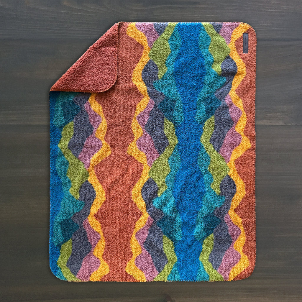 The Sherpa Fleece Waterproof Blanket, a rectangular non-toxic pet blanket with one corner folded, features a vibrant wavy pattern in blue, green, yellow, orange, purple, and pink on a dark background atop a dark wooden surface.