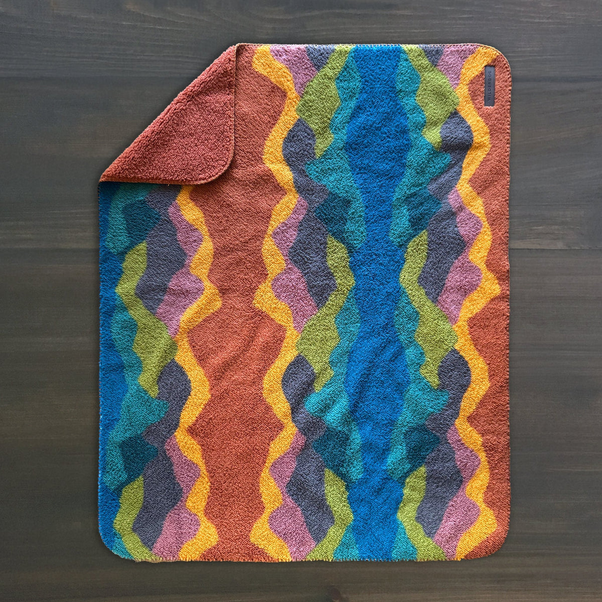 The Sherpa Fleece Waterproof Blanket, a rectangular non-toxic pet blanket with one corner folded, features a vibrant wavy pattern in blue, green, yellow, orange, purple, and pink on a dark background atop a dark wooden surface.
