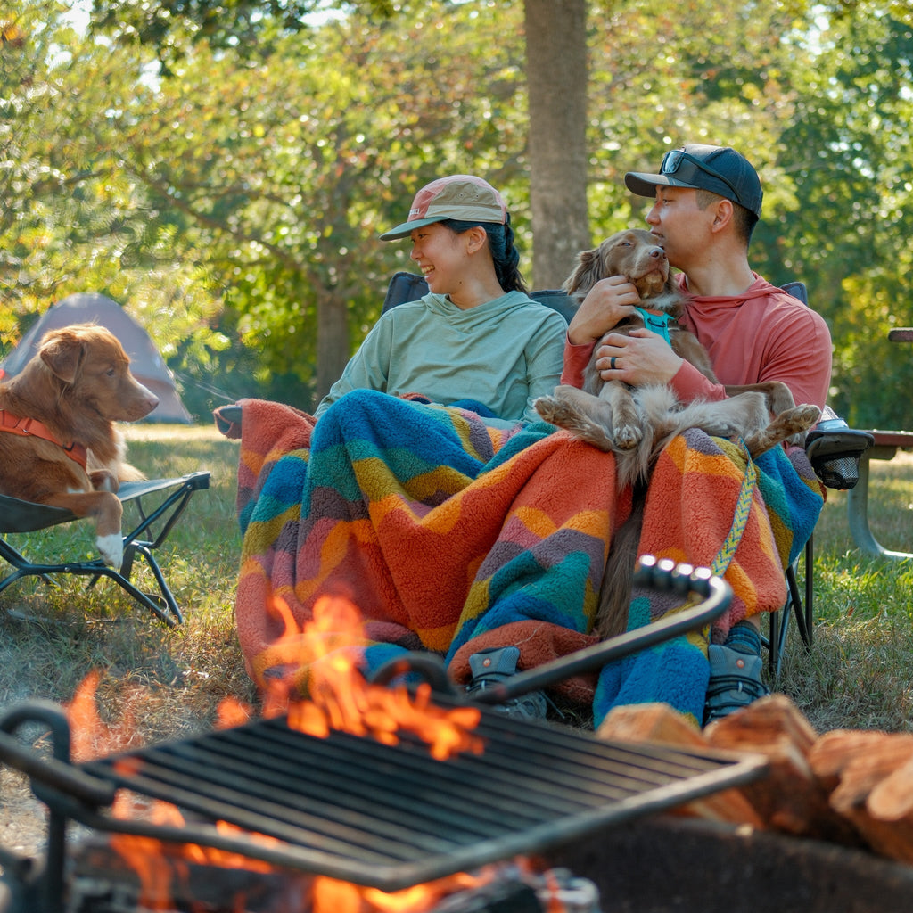 A couple sits in camping chairs by a fire pit, smiling as they share a Sherpa Fleece Waterproof Blanket. One holds a dog on their lap while another dog sits nearby, surrounded by trees and sunlight.