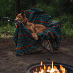 A dog with reddish fur is comfortably curled up on a camping chair covered with a colorful Sherpa Fleece Waterproof Blanket. The chair is placed outdoors on a dirt ground. In the foreground, there is a small fire burning in a fire pit, with a forested background.