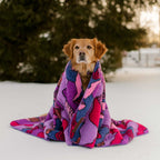 A golden brown dog sits on snow, wrapped in a Sherpa Fleece Waterproof Blanket with pink, purple, and blue tones. Pine trees are visible in the background.