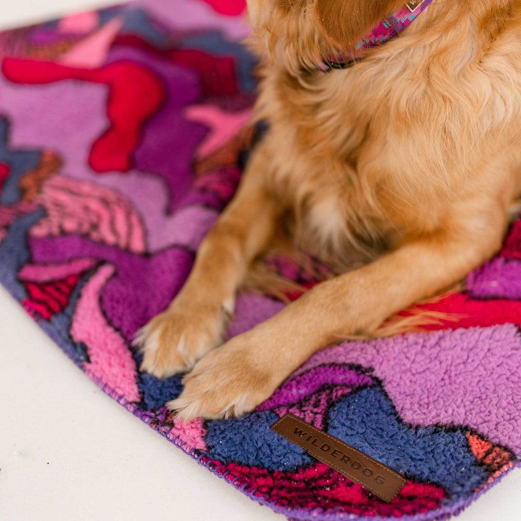 A golden dog rests on the Sherpa Fleece Waterproof Blanket, featuring a vibrant abstract pattern in pink, purple, and red. The non-toxic blanket includes a brown leather “Wilderdog” tag; the dog's front legs and collar are also visible.