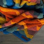 A colorful Sherpa Fleece Waterproof Blanket with an abstract patchwork in orange, blue, teal, yellow, and maroon rests on a wooden surface. A small leather tag reads "WILDERDOG.