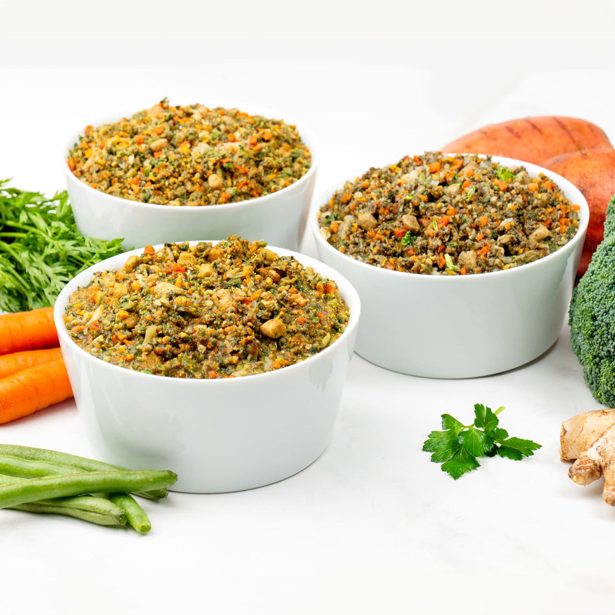 Three white bowls of finely chopped vegetable and meat mix surrounded by fresh carrots, broccoli, green beans, sweet potato, parsley, and ginger