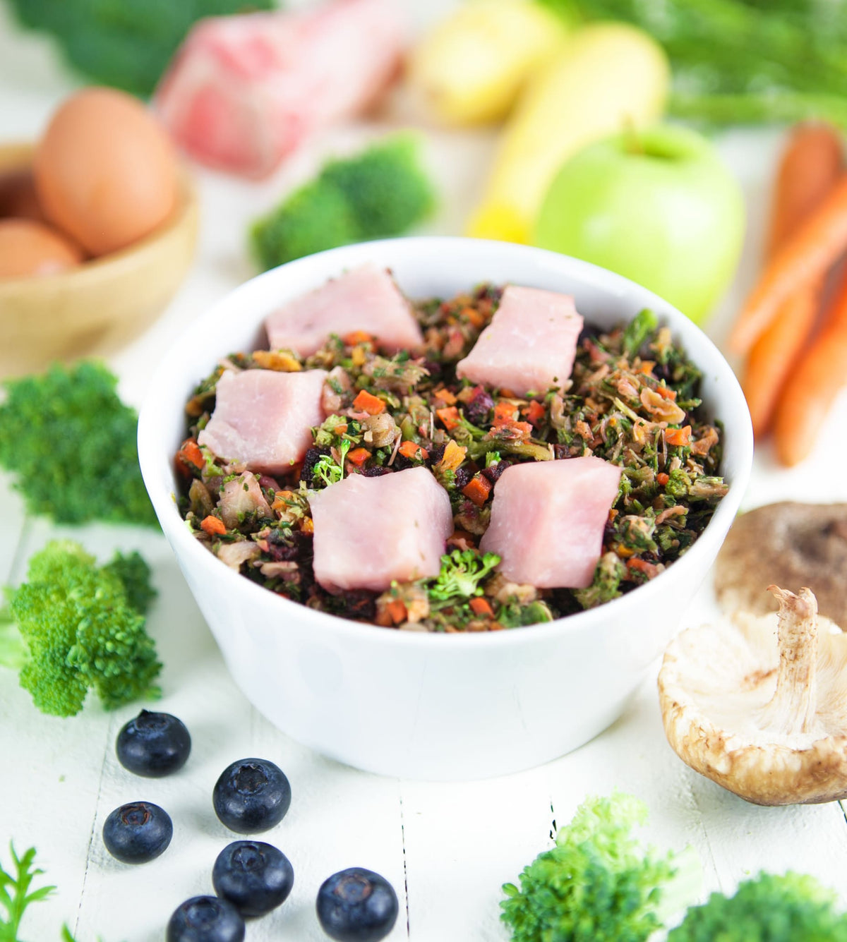 White bowl of fresh raw dog food with meat chunks, vegetables, and herbs, surrounded by whole ingredients