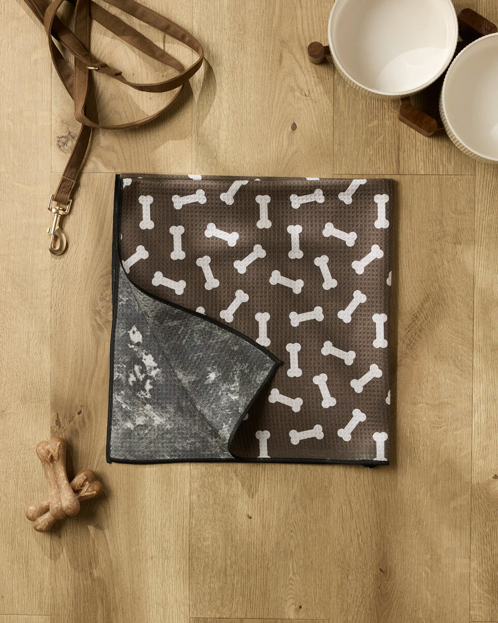 A brown dog mat with white bone patterns from Geometry's Sweet Treats collection lies on a wooden floor, joined by a recycled dog towel, leash, two ceramic bowls on a wooden stand, and two dog bone toys for a cozy corner.