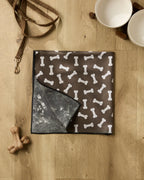 A brown dog mat with white bone patterns from Geometry's Sweet Treats collection lies on a wooden floor, joined by a recycled dog towel, leash, two ceramic bowls on a wooden stand, and two dog bone toys for a cozy corner.