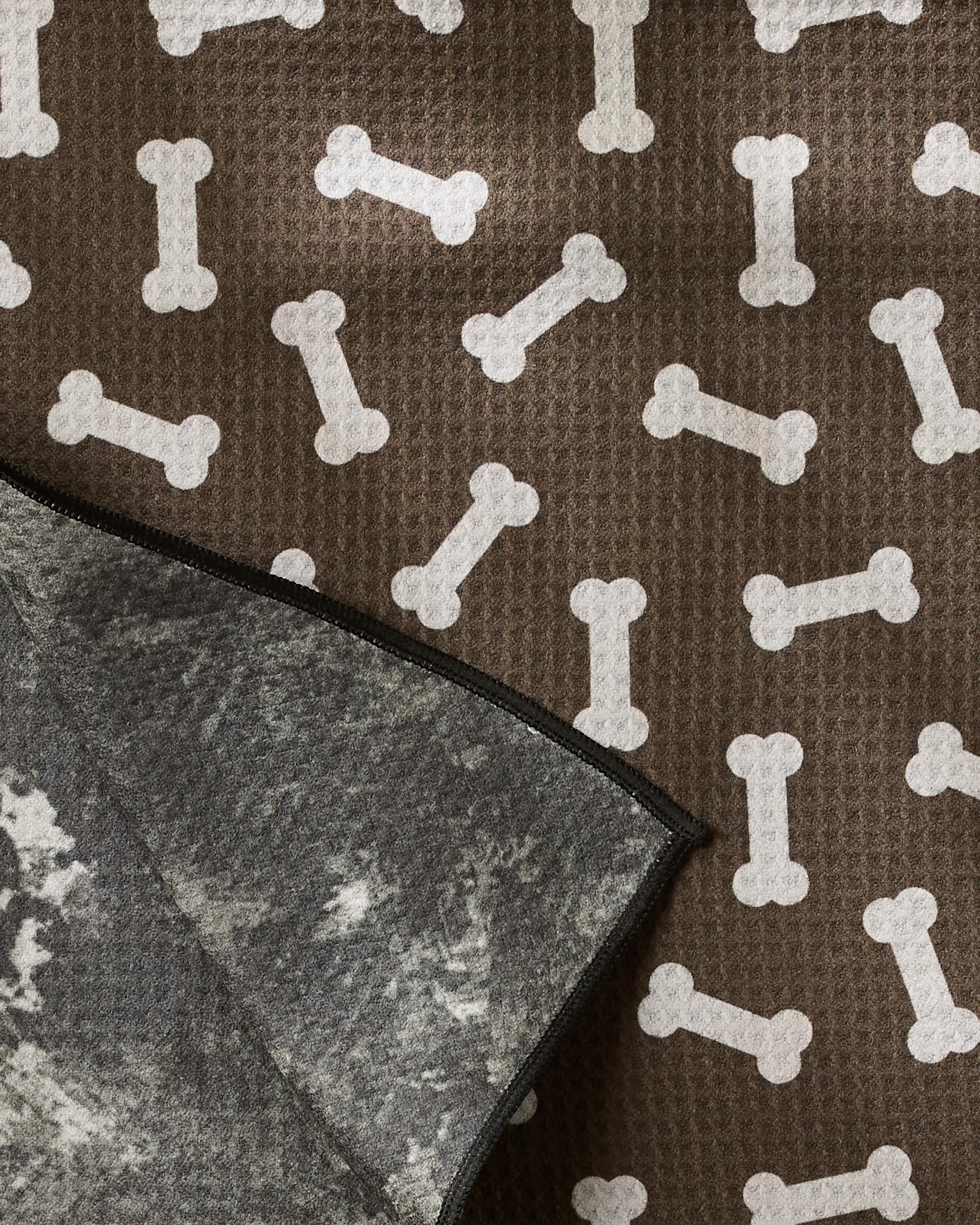 The Geometry Sweet Treats dog towel is brown with white bone patterns and features a gray, mottled underside visible at one folded corner.