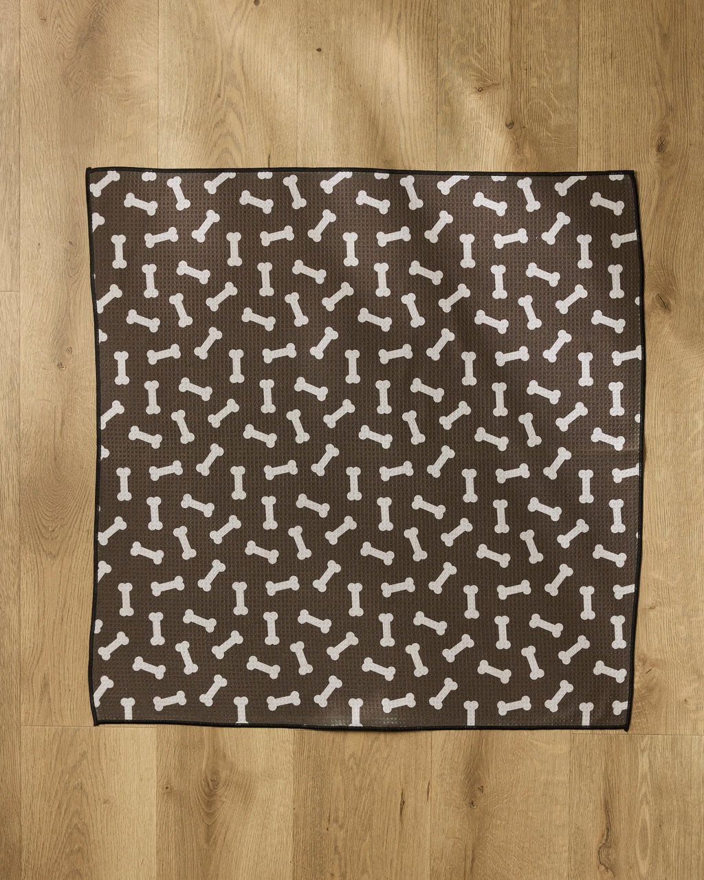 The Geometry Sweet Treats mat is a brown, square dog mat featuring a white bone pattern, made from absorbent towel material, and placed on a wooden floor.