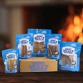 Five blue packages of Best Bully Sticks surround a branded box labeled Hickory Smoked Sampler Box, with a blurred fireplace in the background—ideal for fans of Bully Sticks and Hickory Smoked Dog Chews.