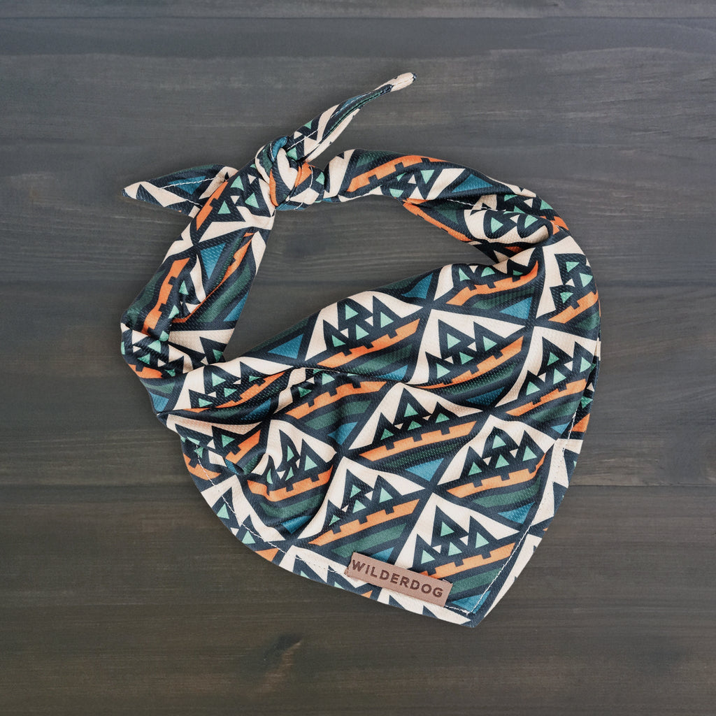 The Camp Bandana is a lightweight dog bandana featuring a blue, orange, and white geometric mountain pattern, tied at the top and labeled "Wilderdog," shown on a dark wooden surface.