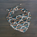 The Camp Bandana is a lightweight dog bandana featuring a blue, orange, and white geometric mountain pattern, tied at the top and labeled "Wilderdog," shown on a dark wooden surface.
