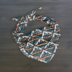The Camp Bandana is a lightweight dog bandana featuring a blue, orange, and white geometric mountain pattern, tied at the top and labeled "Wilderdog," shown on a dark wooden surface.