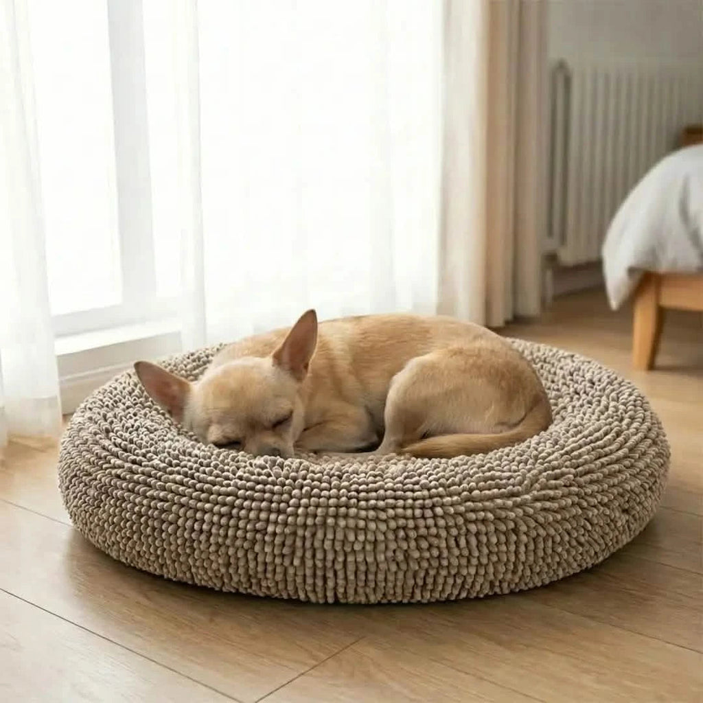 Angled top view of beige round donut dog bed showcasing plush chenille texture on hardwood floor with a sleeping small tan dog