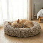 Angled top view of beige round donut dog bed showcasing plush chenille texture on hardwood floor with a sleeping small tan dog