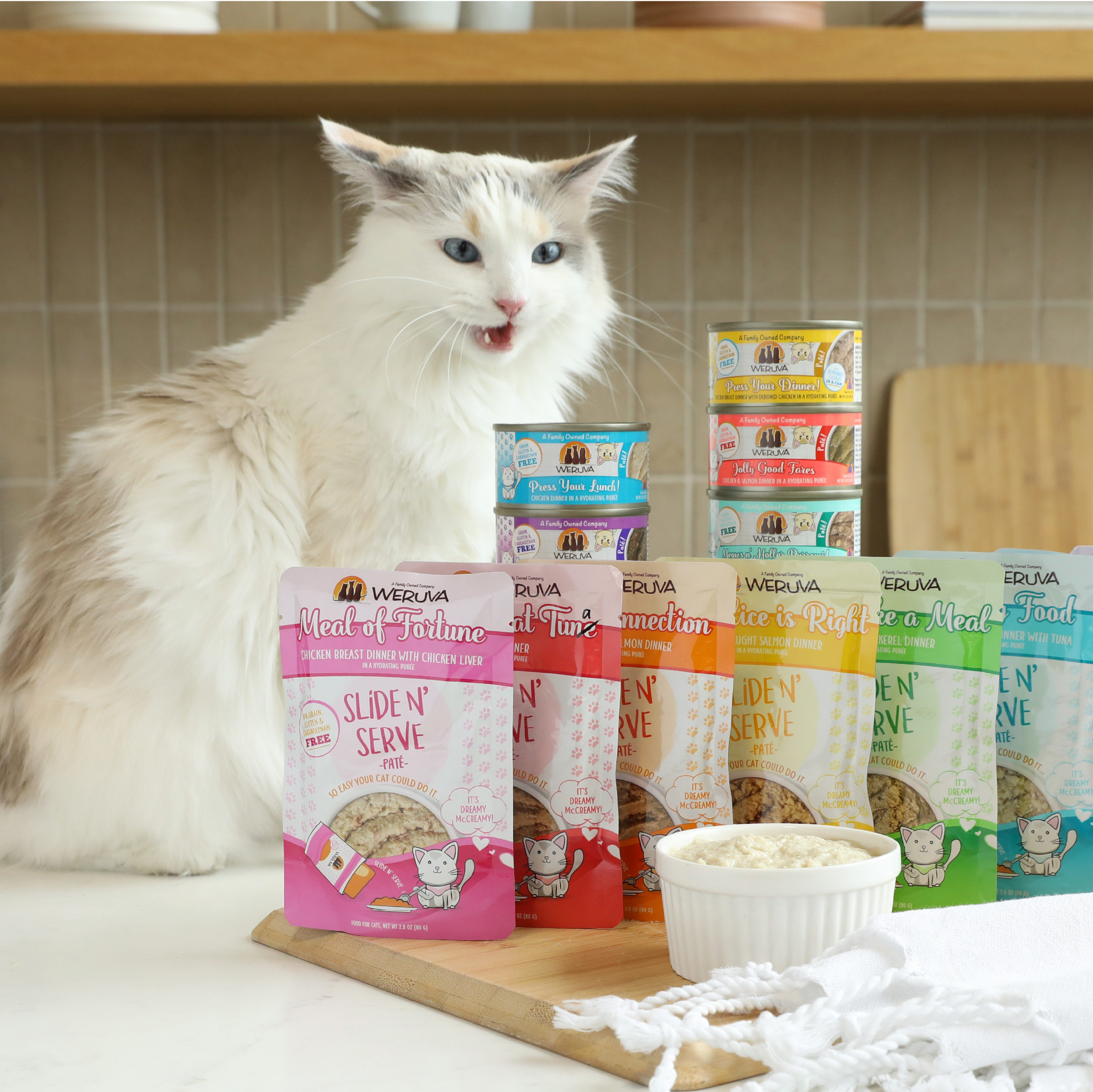 A fluffy white and gray cat sits on a kitchen counter beside stacked Weruva cans and pouches, savoring a bowl of food. It's clearly content, tongue out, enjoying The Suppertime Sweepstakes meals from Weruva.