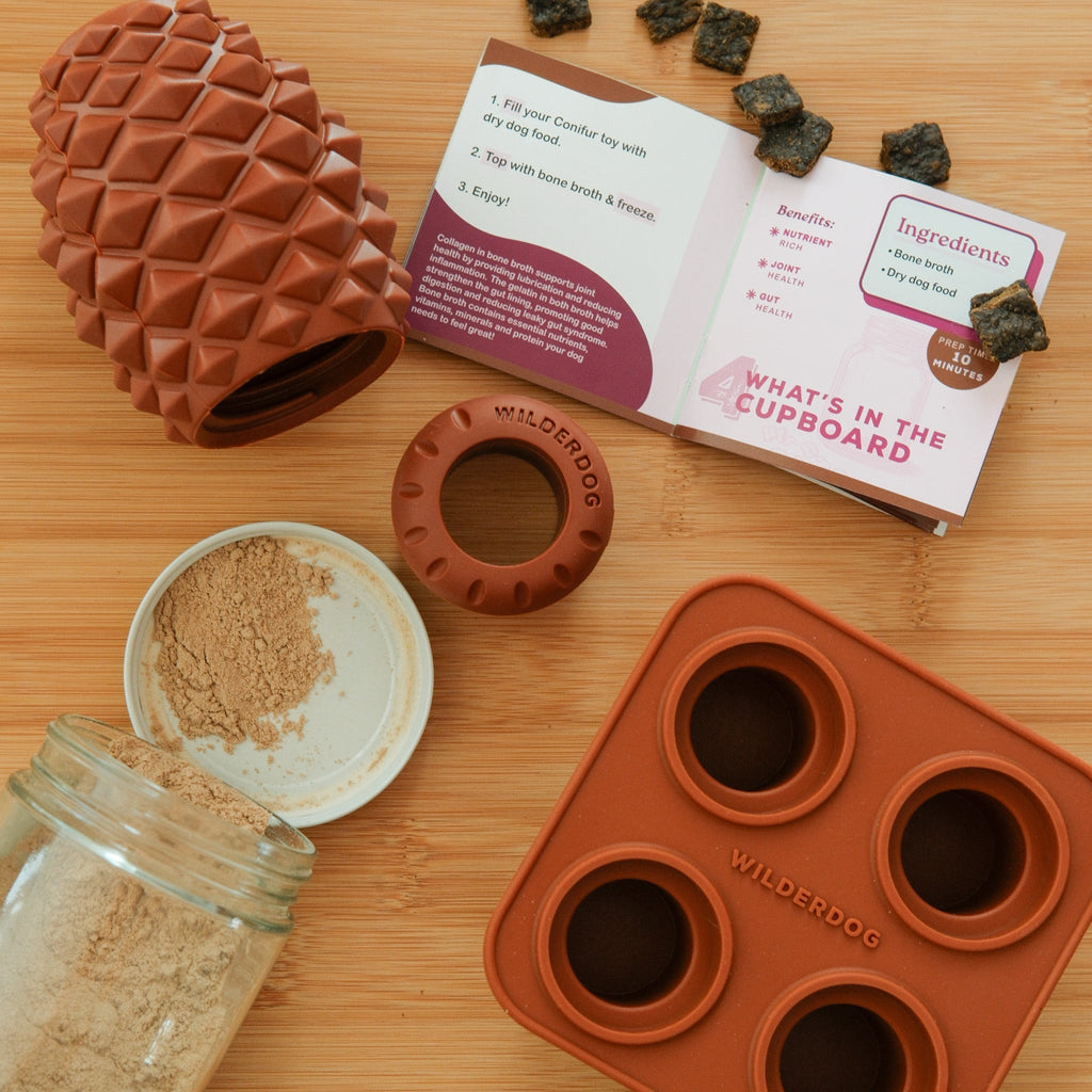 The Conifur set includes a silicone pinecone-shaped dog toy, recipe booklet, 4 brown dog treats, a powder jar, and a silicone treat mold—all Wilderdog branded and arranged on wood.