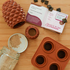 The Conifur set includes a silicone pinecone-shaped dog toy, recipe booklet, 4 brown dog treats, a powder jar, and a silicone treat mold—all Wilderdog branded and arranged on wood.