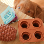 A brown dog looks up at a table with The Conifur silicone treat mold, a textured rubber toy, and an open Wilderdog Consfur Recipe Book.
