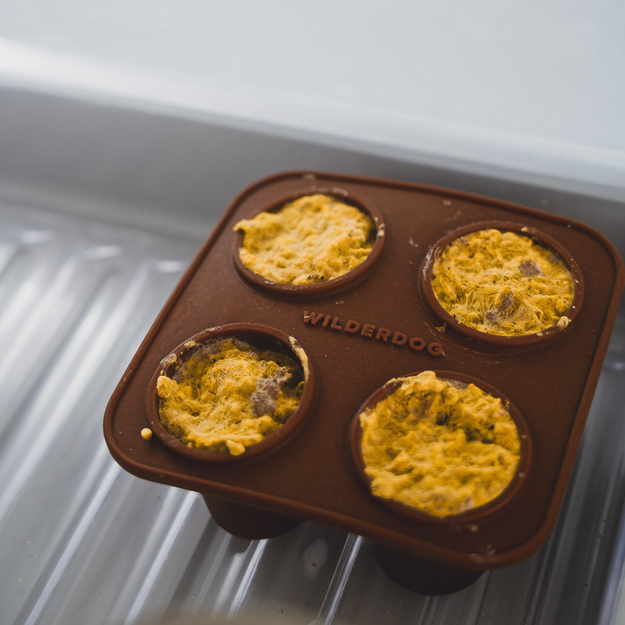 The Conifur is a brown silicone mold with "WILDERDOG" branding, holding four round portions of yellow dog treats, perfect as a creative frozen enrichment toy for your pup.