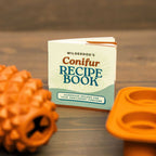 A small recipe book titled "Wilderdog's Conifur Recipe Book" stands upright next to The Conifur—an orange pinecone-shaped dog toy—and an orange silicone mold tray on a wooden surface.