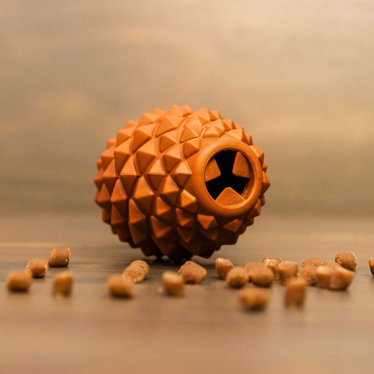 The Conifur, a textured brown rubber dog toy with a star-shaped hole, rests on a wooden surface surrounded by scattered square dog treats.